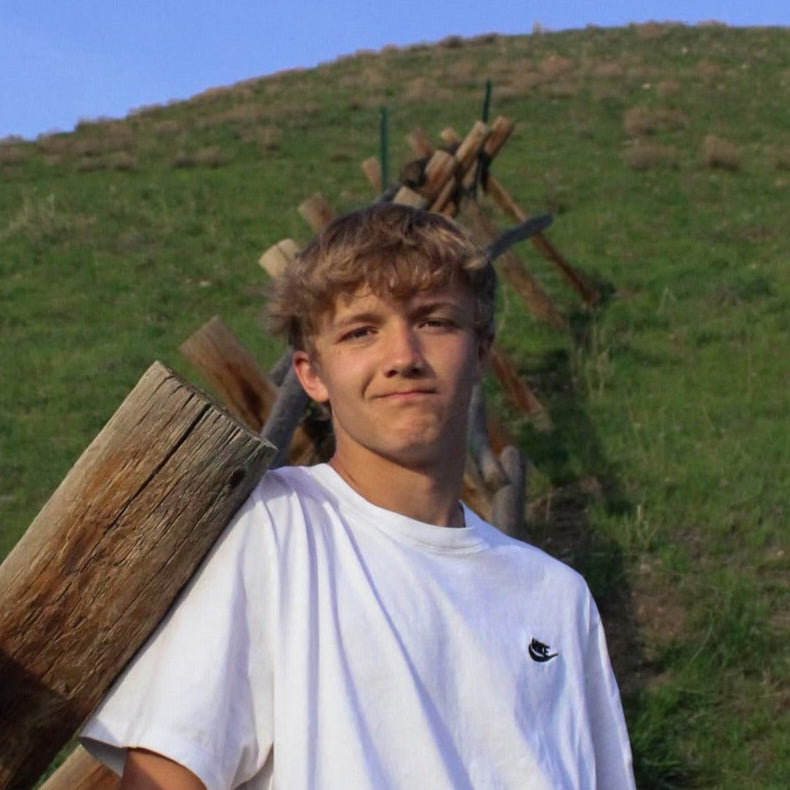 Person leaning against a wooden fence with a grassy hill and blue sky in the background